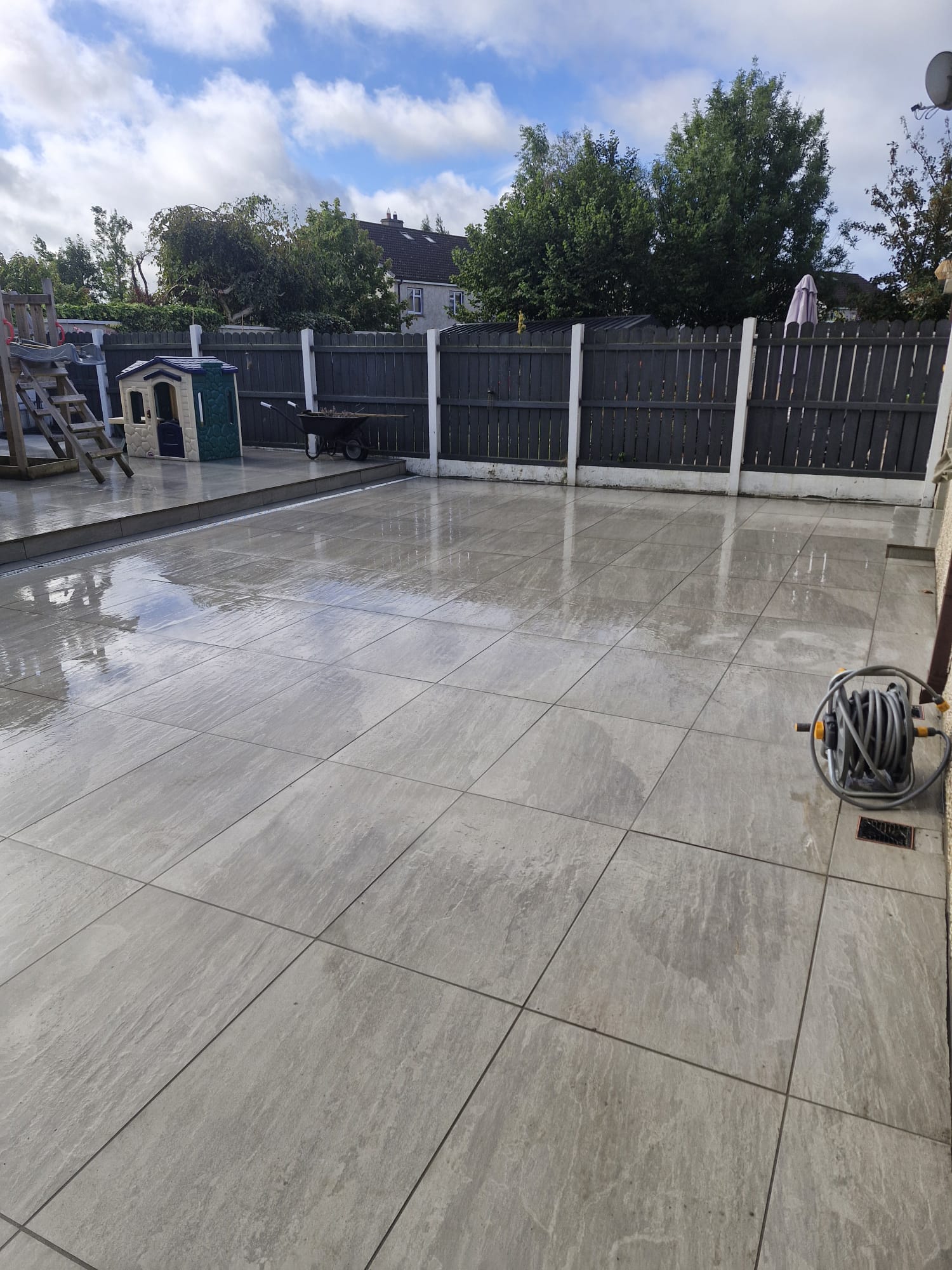 Wide grey porcelain patio filling a back garden beside dark timber fencing | Farrell Exteriors